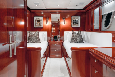 Two neatly made twin beds with decorative pillows are in the foreground, while the background reveals a luxurious cabin with polished wood paneling and oval windows.