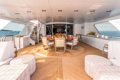 Plush white seating arrangements in the foreground lead to a circular dining table set for a meal on a yacht's deck, with a calm ocean visible through large windows in the background.