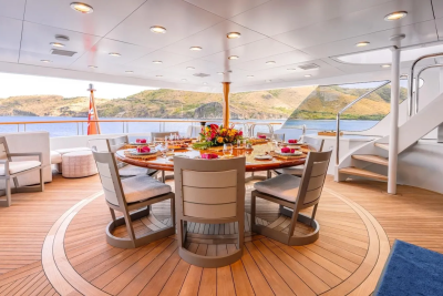 A circular dining table set for a meal on a yacht's deck is surrounded by chairs, with a scenic view of a bay and hills in the background.