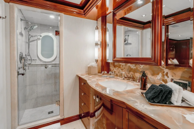 A luxurious yacht bathroom features a marble vanity with a sink and towels in the foreground, and a glass-enclosed shower with an oval mirror in the background.