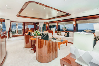 Luxurious cream carpets and polished wood furniture fill the foreground of a yacht's elegant lounge, while large windows in the background reveal a serene ocean view.