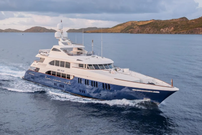 A large blue and white yacht cruises through calm water with a hilly, green coastline in the background.