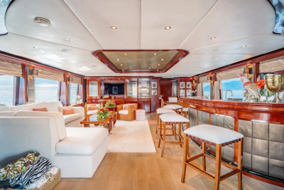 A luxurious yacht interior features a white sectional sofa and coffee table in the foreground, with a polished wood bar and stools in the background, all illuminated by natural light from large windows.