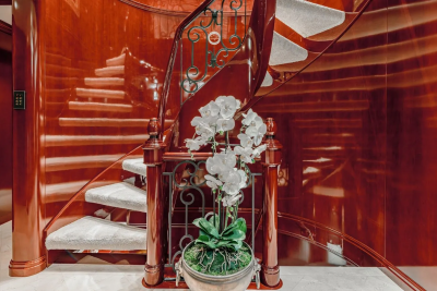 A spiral staircase with white carpeted steps and a polished red wood railing is adorned with a potted orchid in the foreground, while the background features the continuation of the gleaming red wood walls and staircase.