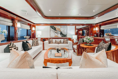 A luxurious yacht interior features plush white sofas and a polished wooden coffee table in the foreground, with large windows revealing a serene ocean view in the background.