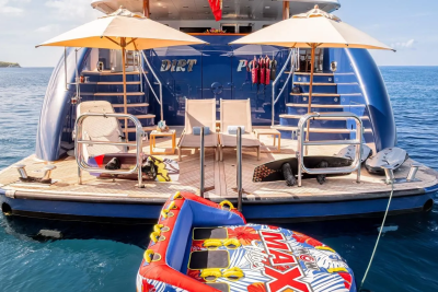 A colorful inflatable tube floats in the foreground in front of a large blue yacht with two umbrellas and lounge chairs on its deck, all set against a calm blue ocean and distant green hills.