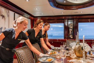 Three women in black uniforms set a formal dining table on a yacht, with the ocean visible through large windows in the background.
