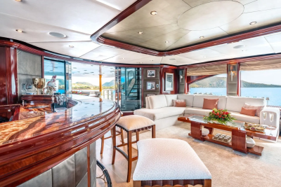 A luxurious yacht interior features a polished wooden bar with stools in the foreground and a spacious lounge area with a sectional sofa and ocean views in the background.