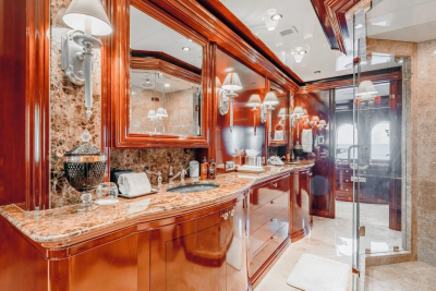 A luxurious bathroom with a marble countertop and polished wood cabinets in the foreground, and a glass-enclosed shower and a doorway leading to another room in the background.