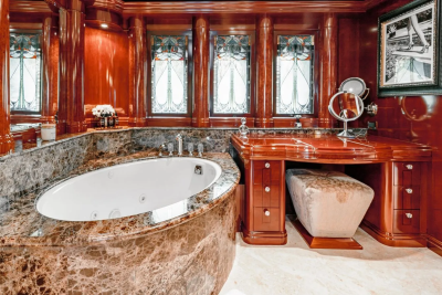 A luxurious bathroom features a marble-encased jacuzzi in the foreground and a polished wood vanity with a mirror and stool in the background, all set against ornate wooden paneling and stained-glass windows.