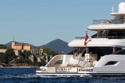 A large white yacht with a red flag sails past a historic stone building on a hill overlooking the sea.