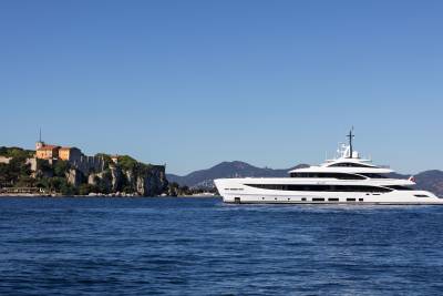A large white yacht sails on calm blue water in the foreground, with a historic stone building on a cliff and distant mountains in the background under a clear blue sky.