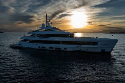 A large white yacht floats on calm water in the foreground, with a dramatic sunset casting a warm glow over distant mountains in the background.