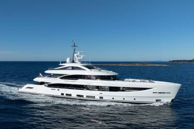 A sleek white yacht cruises through calm blue waters with a distant, tree-lined coastline under a clear sky.