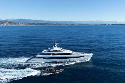 A large white yacht cruises through calm blue water, leaving a foamy wake, with a smaller tender boat trailing behind it, and a hazy coastline with mountains in the distance under a clear sky.