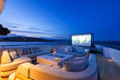A luxurious yacht deck with comfortable seating and a large outdoor screen is set against a twilight sky and a distant coastline.