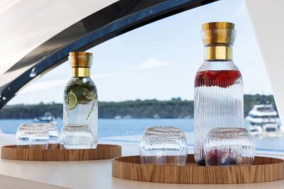 Two carafes of infused water and glasses sit on wooden trays in the foreground, with a blurred seascape and yachts in the background.