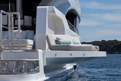 A luxurious yacht's extended deck with plush seating is anchored in calm blue water with a tree-lined coastline in the distance.