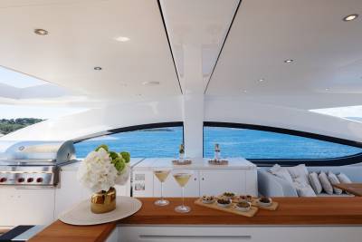 A wooden bar with cocktails and appetizers in the foreground is set against a backdrop of the ocean viewed through a large window on a modern yacht.