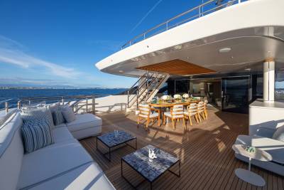 A luxurious yacht deck with a white sectional sofa and patterned coffee tables in the foreground, and a dining table set for a meal under a covered area, with the ocean and distant coastline in the background.