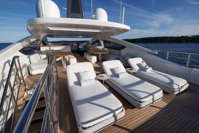 Three white lounge chairs with blue pillows sit on the wooden deck of a modern yacht in the foreground, with a calm blue ocean and a clear sky in the background.