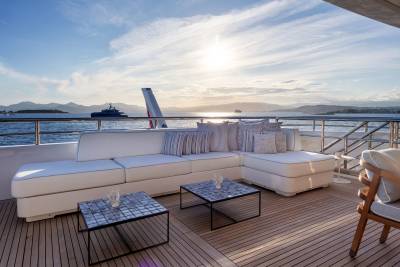 A white sectional sofa and two coffee tables sit on a wooden deck in the foreground, with a calm sea, distant islands, and a bright sky in the background.