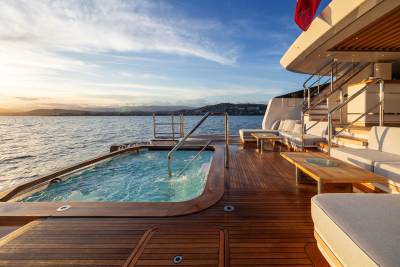 A bubbling jacuzzi and plush seating area on a yacht's deck overlook a calm sea and distant coastline under a soft sunset sky.