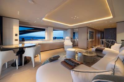 A modern yacht interior features a circular dining table in the foreground and a spacious, well-lit living area with large windows overlooking the ocean in the background.