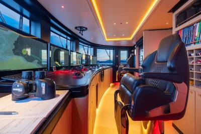 In the foreground, a pair of binoculars rests on a chart, while the background reveals a modern yacht bridge with multiple navigation screens and comfortable seating.