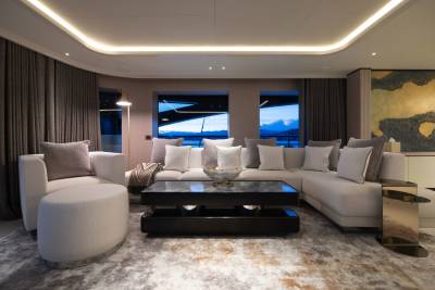 A plush sectional sofa and ottoman fill the foreground of a luxurious yacht lounge, while large windows in the background reveal a serene ocean sunset.