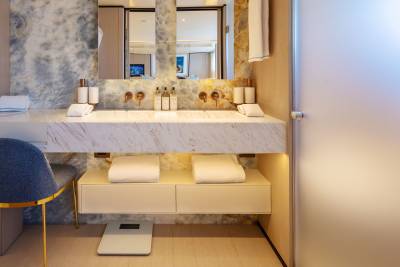 A modern bathroom vanity with a marble countertop and dual sinks is in the foreground, while a frosted glass door and a reflection of a hotel room are in the background.