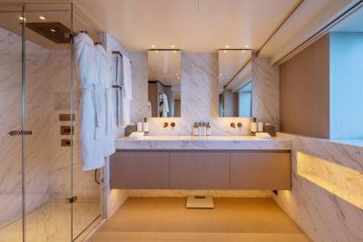 Plush white robes hang in a glass-enclosed shower in the foreground, while a sleek double vanity with marble accents and illuminated cabinetry fills the background.