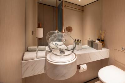 A modern bathroom features a clear glass vessel sink on a marble countertop in the foreground, with a toilet and toiletries visible in the background.