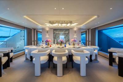 A luxurious dining room with a set table and plush chairs in the foreground is framed by large windows offering views of the ocean and sky in the background.