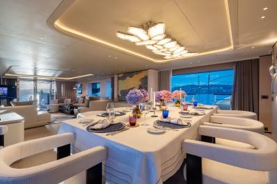 A formal dining table set with colorful glasses and floral centerpieces is in the foreground, while a spacious, modern living area with large windows overlooking the ocean is in the background.