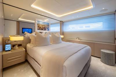 A luxurious yacht bedroom features a plush king-sized bed in the foreground and a large window with blinds and a television in the background.