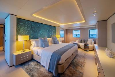 A luxurious yacht bedroom with a large bed in the foreground and a seating area with windows overlooking the water in the background.