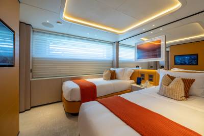 Two beds with orange throws are in the foreground, while a large window with blinds and a mounted television are in the background.