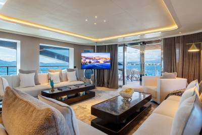 Plush white sofas and modern coffee tables fill the foreground of a luxurious yacht's living room, while large windows reveal a serene ocean view and an outdoor seating area in the background.