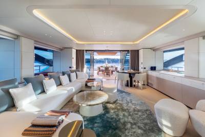A luxurious yacht interior with a plush white sectional sofa and modern coffee tables in the foreground, opening to a sun-drenched dining area and a serene waterfront view in the background.
