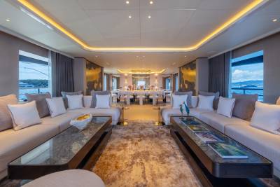Plush sofas and modern coffee tables fill the foreground of a luxurious yacht's lounge, while a long dining table and illuminated artwork adorn the background.