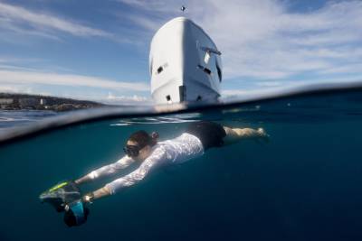 A woman swims underwater with a sea scooter in the foreground, while the hull of a white boat is visible above the waterline in the background.