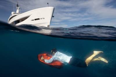 A man in a suit swims underwater on a red scooter, with a large white yacht sailing above him in the blue ocean.