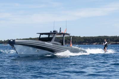 A sleek, dark gray speedboat cuts through blue water, leaving a white wake as a person on a wakeboard follows behind, with a tree-lined coast visible in the distance under a clear sky.