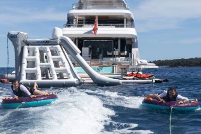 Two people ride inflatable tubes through choppy blue water in front of a large white yacht with an inflatable water slide attached.