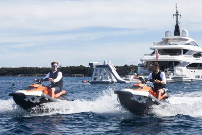 Two men ride jet skis in the foreground, leaving wakes in the water, while a large yacht and a floating water slide are visible in the background.
