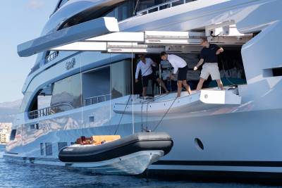 A small inflatable boat is being lowered from a large white yacht, with a few people on deck and buildings visible in the background.