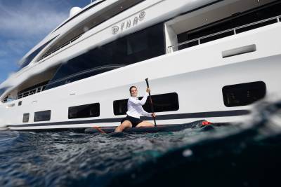 A woman paddles a stand-up paddleboard in the foreground, with a large white yacht named "Dyna" in the background.