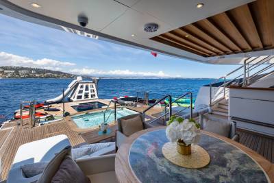 A luxurious yacht deck with a dining table in the foreground and a pool, waterslide, and ocean in the background.
