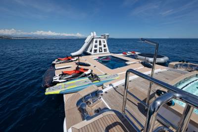 A luxurious yacht deck in the foreground holds jet skis and paddleboards, while a floating water park with a slide and pool is anchored in the calm blue sea under a clear sky in the background.